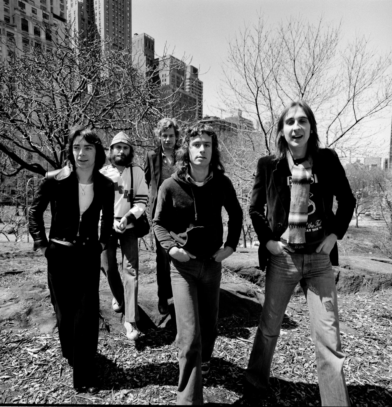 Pięciu młodych mężczyzn w casualowych ubraniach spaceruje przez park na tle wysokich budynków, drzewa wokół są bez liści, a atmosfera jest swobodna i wiosenna.