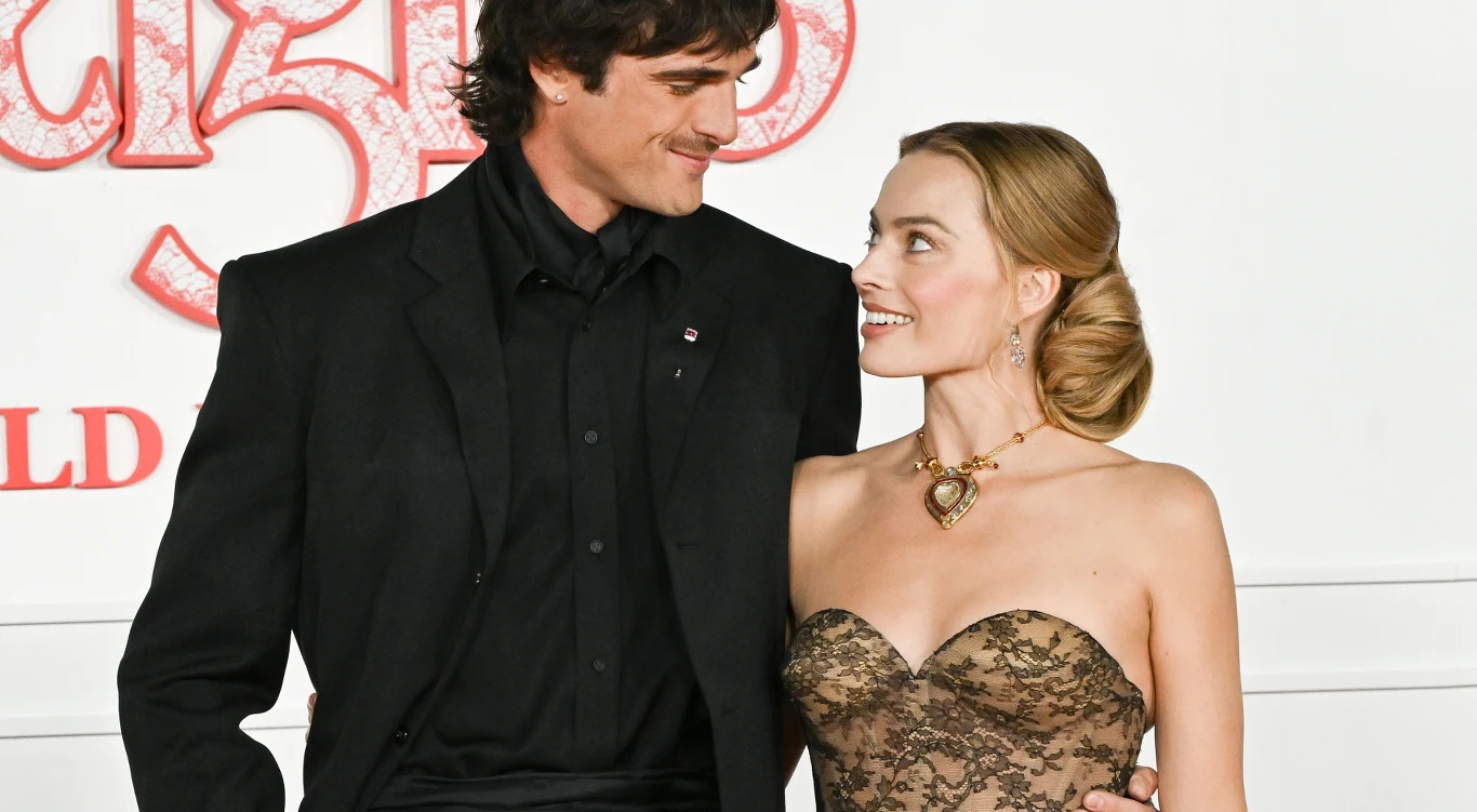 Jacob Elordi i Margot Robbie na światowej premierze filmu "Wichrowe wzgórza" w Hollywood, 28 stycznia 2026 Elegancko ubrany mężczyzna w czarnym garniturze stoi obok kobiety w stroju z koronkową górą i złotym naszyjnikiem. Oboje patrzą na siebie z uśmiechem, tworząc przyjazną i ciepłą atmosferę. W tle widoczne białe i czerwone elementy dekoracyjne.