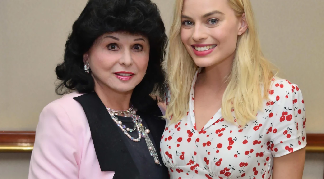 Yola Czaderska-Hayek i Margot Robbie Dwie kobiety stoją obok siebie i uśmiechają się do kamery, jedna ubrana w jasną marynarkę i biżuterię, druga w białą bluzkę w wiśnie, obie zadbane i eleganckie.