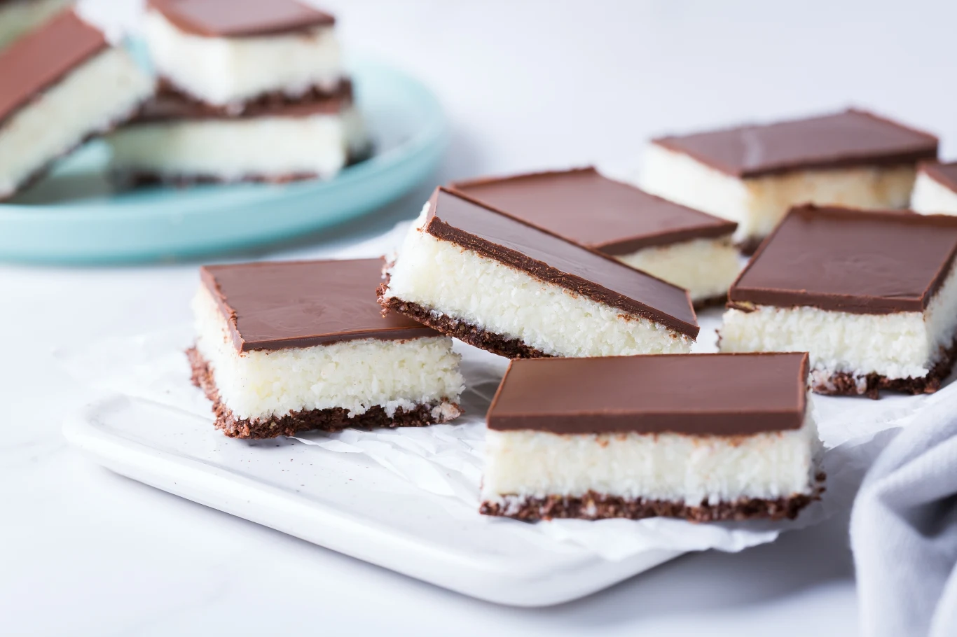 Kwadratowe ciastka z wyraźną warstwą kokosową pokrytą gładką polewą czekoladową, poukładane na papierze do pieczenia. W tle widać więcej ciastek na jasnoniebieskim talerzu.