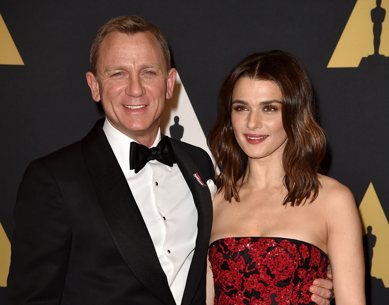 Daniel Craig i Rachel Weisz na rozdaniu Oscarów 2015 Elegancka para w wieczorowych strojach, mężczyzna w czarnym smokingu z muszką, kobieta w czerwonej sukni z czarnym wzorem, uśmiechnięci, pozują na tle ściany z motywem statuetek filmowych.