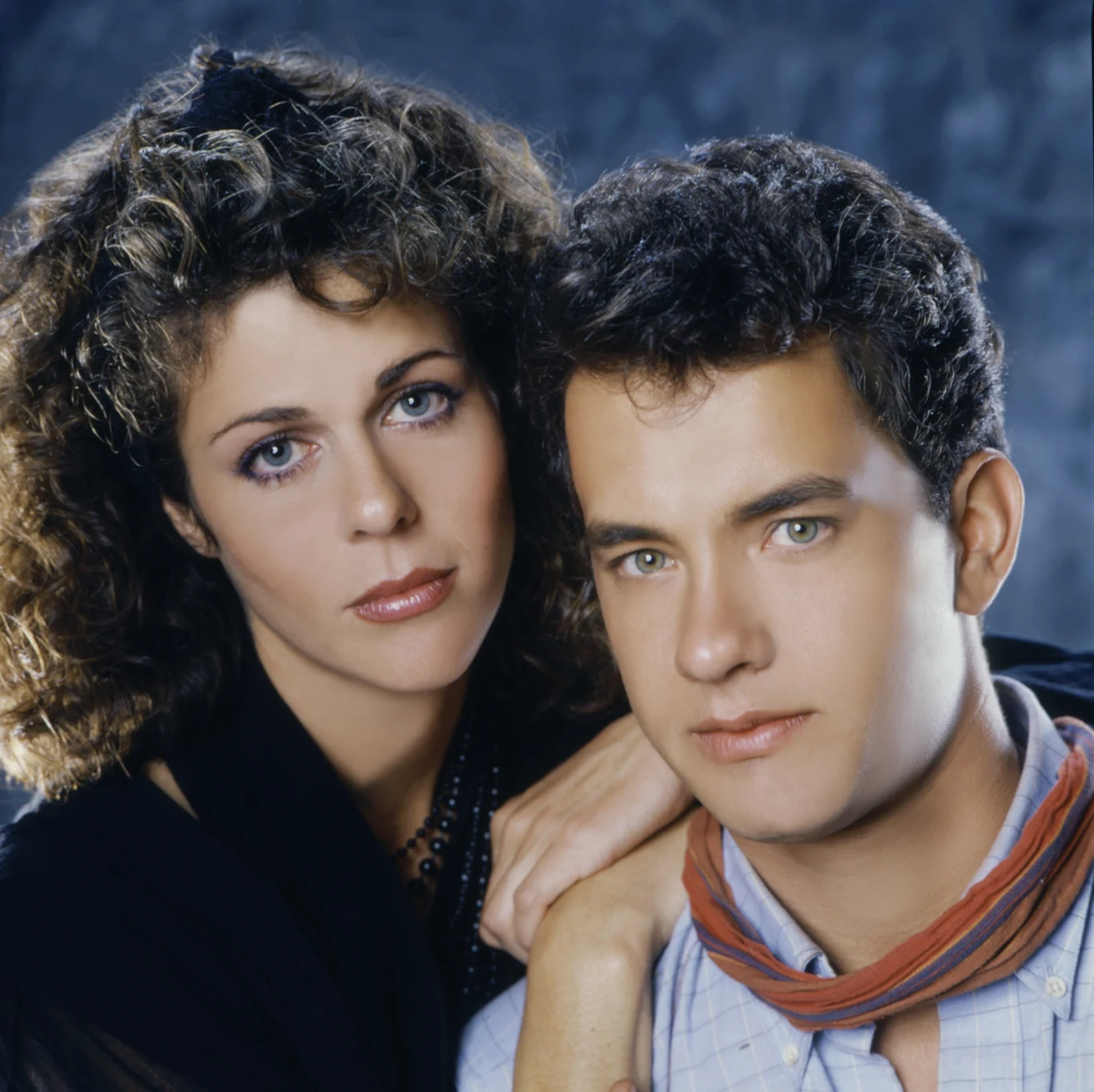 Rita Wilson i Tom Hanks w filmie "Ochotnicy" (1985) Portret młodej kobiety i mężczyzny obejmujących się delikatnie, oboje patrzą prosto w obiektyw, ich twarze są spokojne i wyraziste na jednolitym, ciemnym tle.