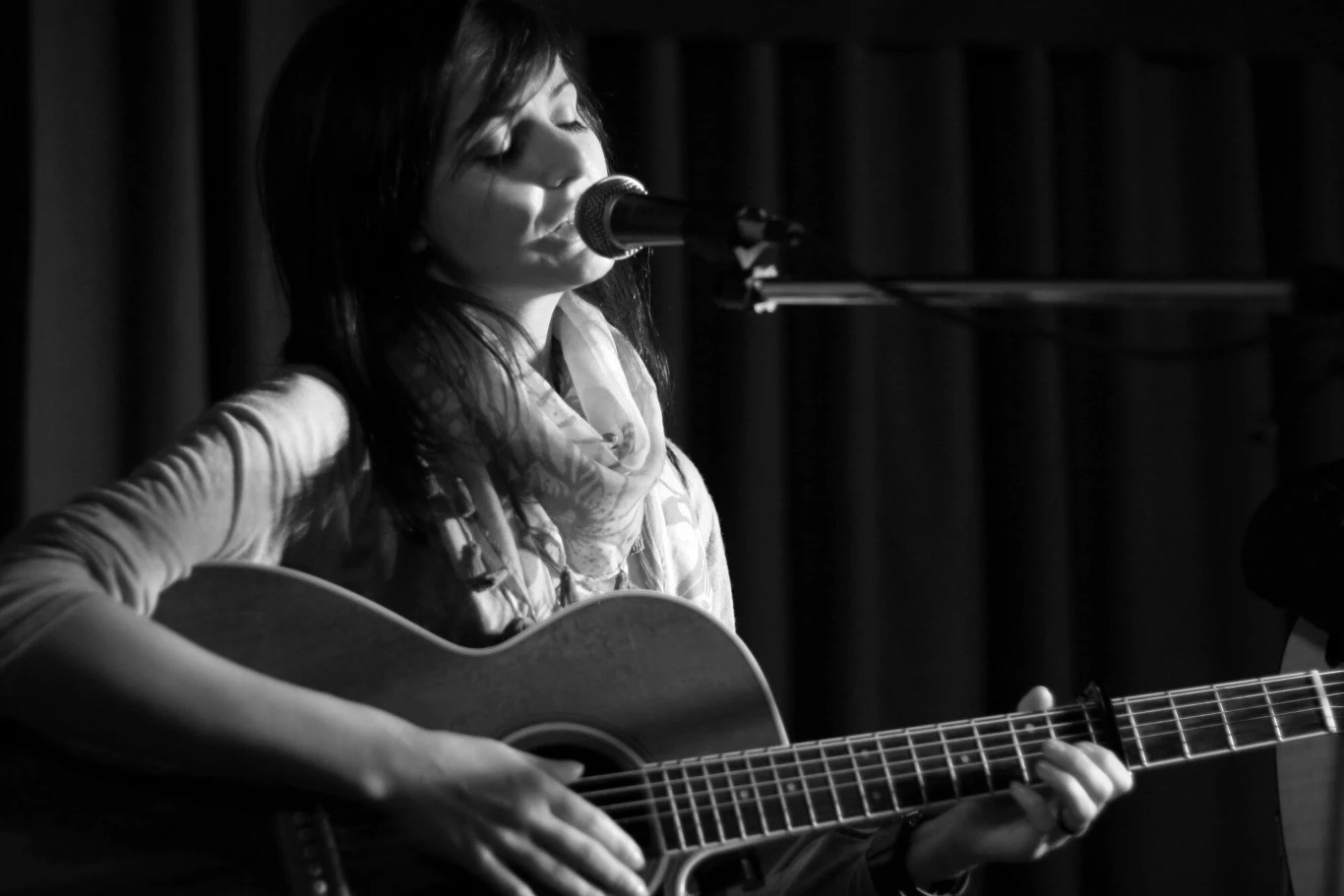 Dominika Żukowska miała 41 lat Kobieta śpiewająca do mikrofonu podczas gry na gitarze akustycznej, skupiona na muzyce, półmrok podkreślający nastrój sceny.