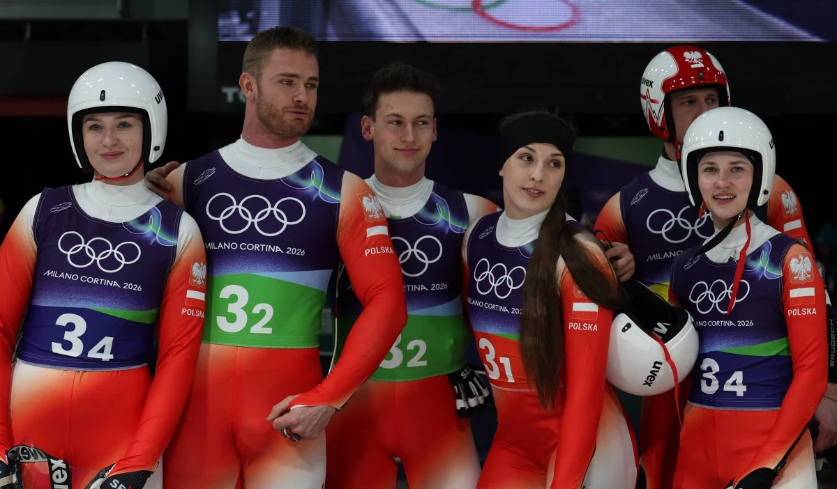 Grupa polskich sportowców ubrana w olimpijskie stroje startowe do saneczkarstwa, stojąca razem na torze. Widoczne numery na ich kombinezonach oraz logotyp igrzysk olimpijskich Milano Cortina 2026.