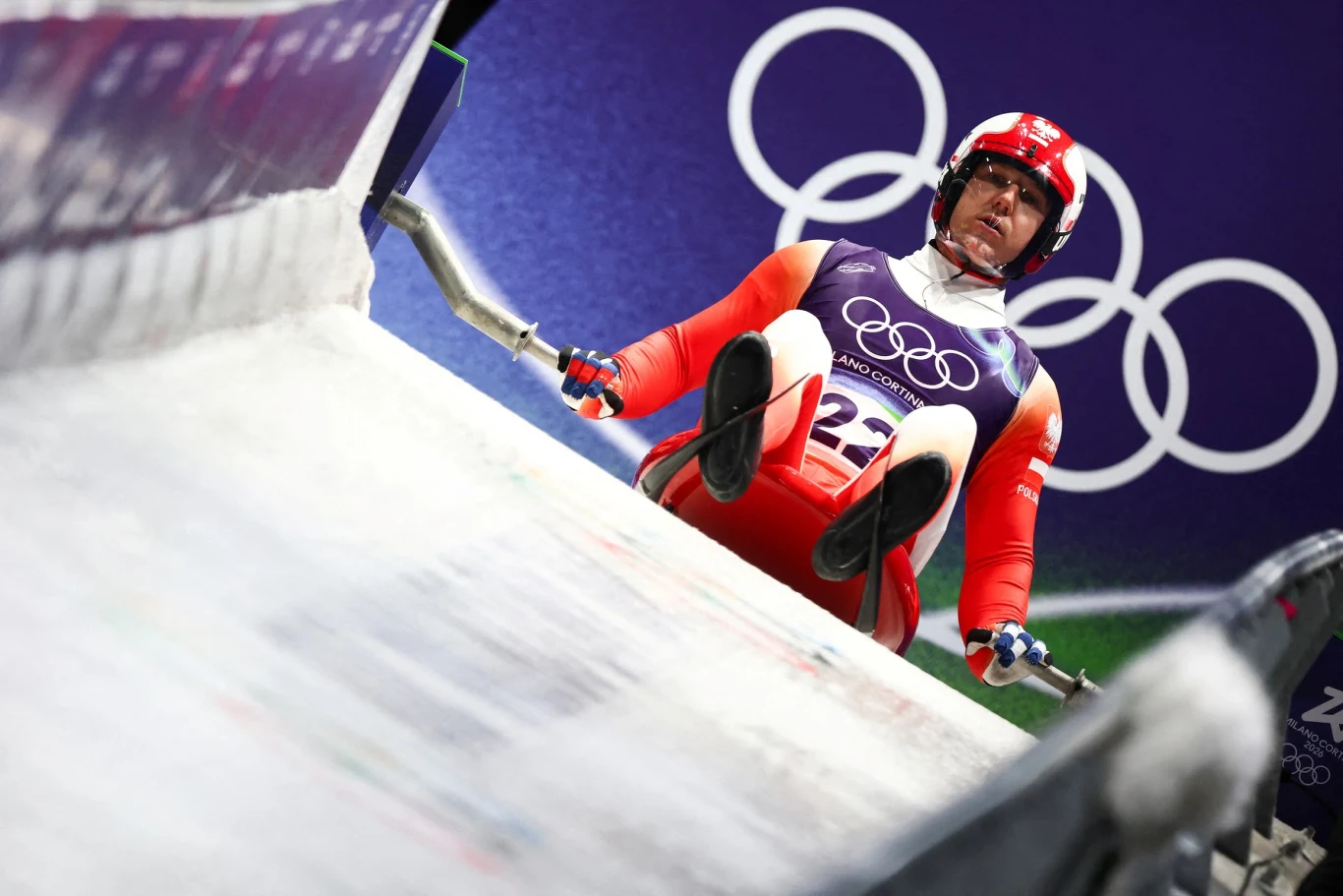 Sportowiec w czerwonym kombinezonie w trakcie startu w zawodach saneczkarskich podczas zimowych igrzysk olimpijskich, w tle widoczne olimpijskie kółka.