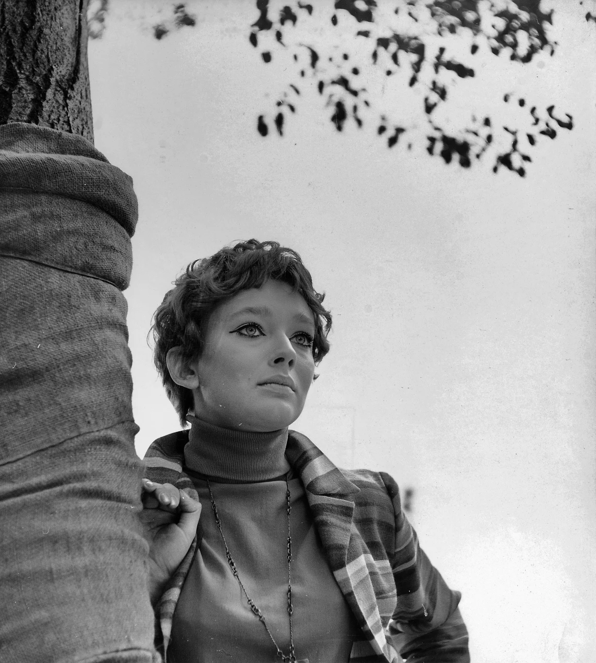 Młoda kobieta z krótkimi, kręconymi włosami oparta o drzewo, ubrana w golf i marynarkę w kratę, patrzy zamyślona w dal. W tle liście drzewa na jasnym, rozmytym niebie.