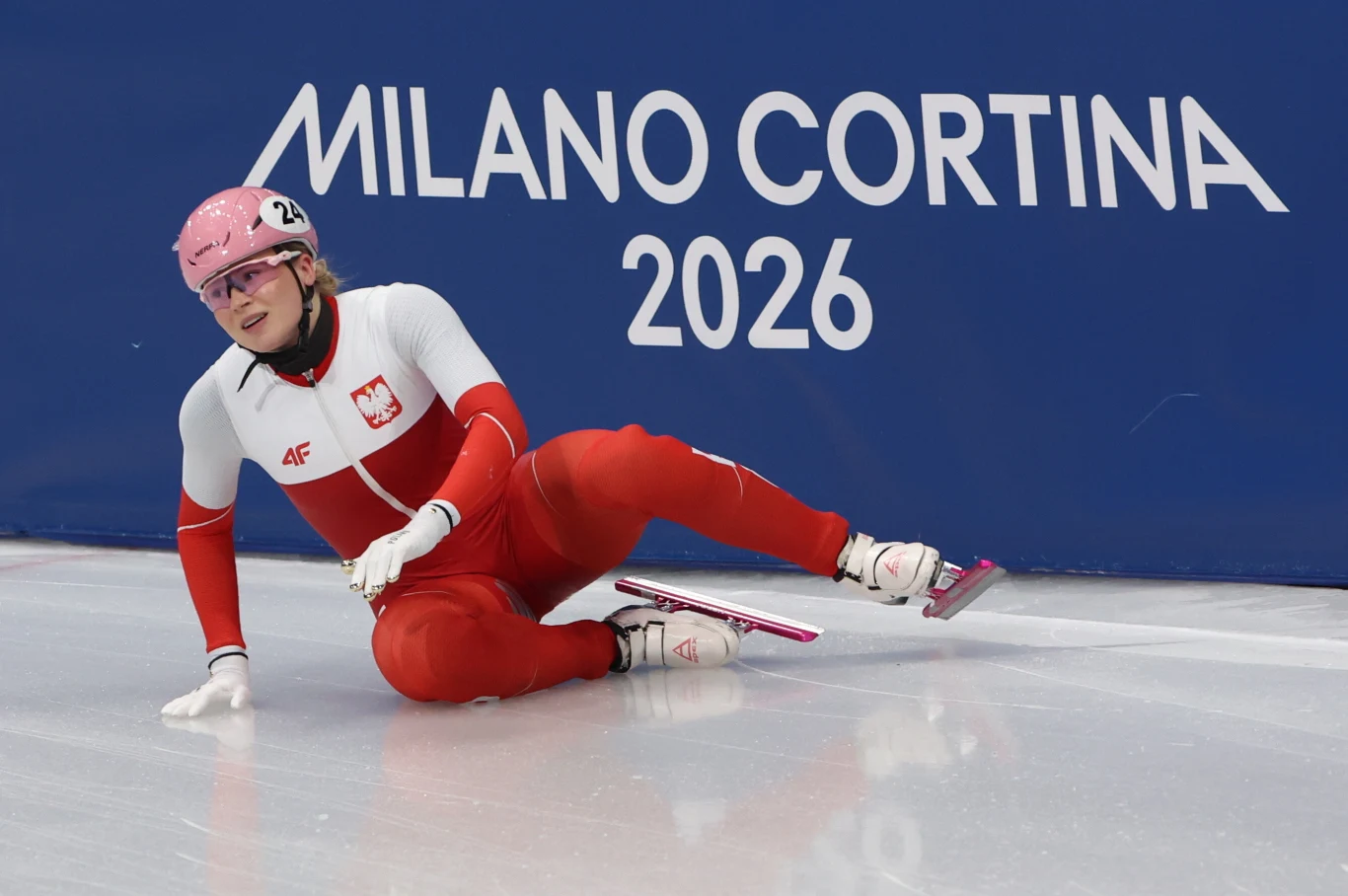 Zawodniczka w stroju reprezentacji Polski siedzi na lodowisku w pozie sugerującej upadek podczas zawodów, w tle widoczny niebieski baner z napisem MILANO CORTINA 2026.