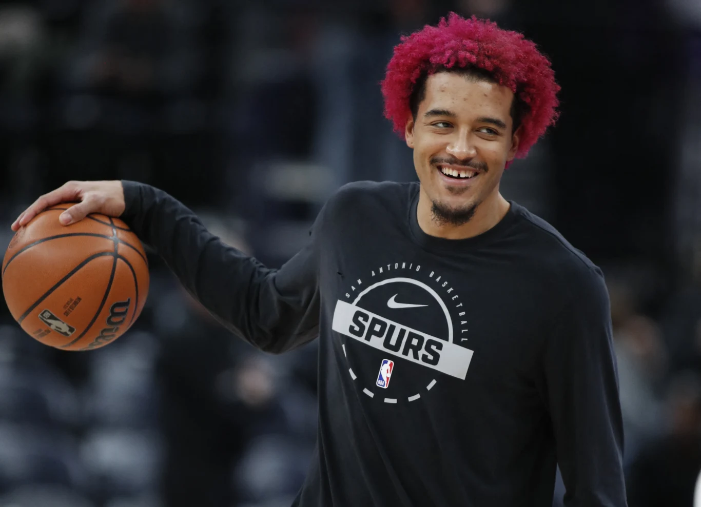 Sportowiec z burzą różowych włosów ubrany w bluzę z logo San Antonio Spurs, trzymający piłkę do koszykówki i uśmiechnięty podczas treningu na hali sportowej.