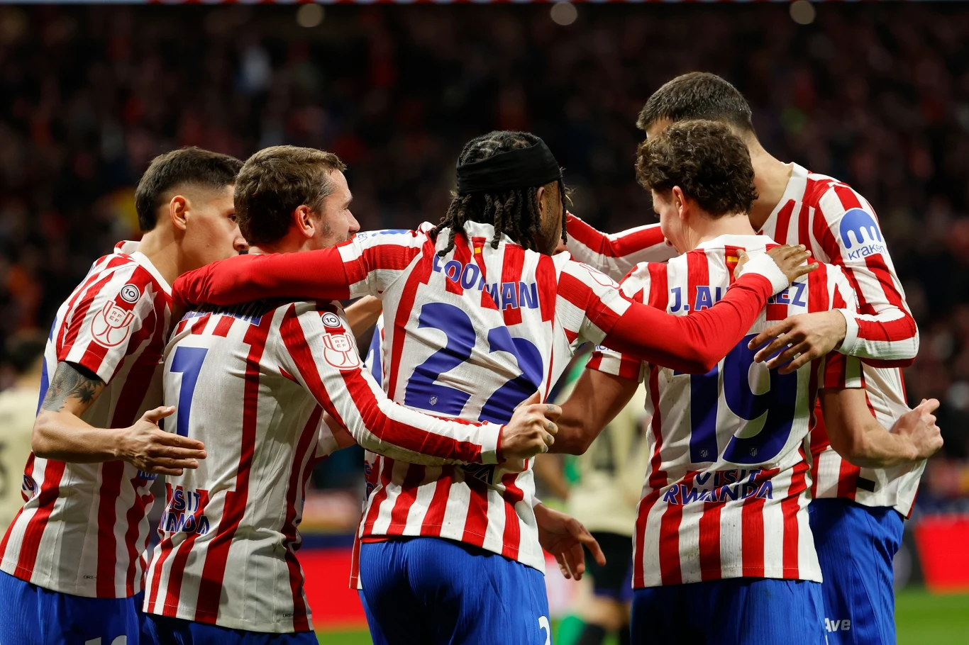 Grupa piłkarzy w czerwono-białych koszulkach świętuje razem na boisku po zdobyciu gola. Zawodnicy obejmują się ramionami i stoją w kręgu, tworząc atmosferę wspólnego triumfu. Tło jest rozmyte, skupiając uwagę na emocjach sportowców.