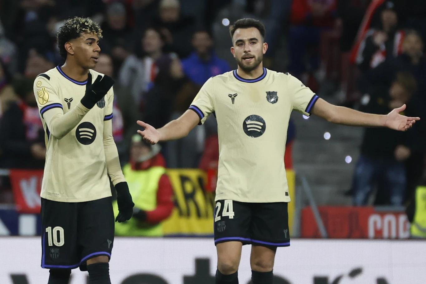 Dwóch piłkarzy w jasnożółtych koszulkach z logo Spotify gestykuluje na boisku podczas meczu, w tle widoczni kibice i flagi.