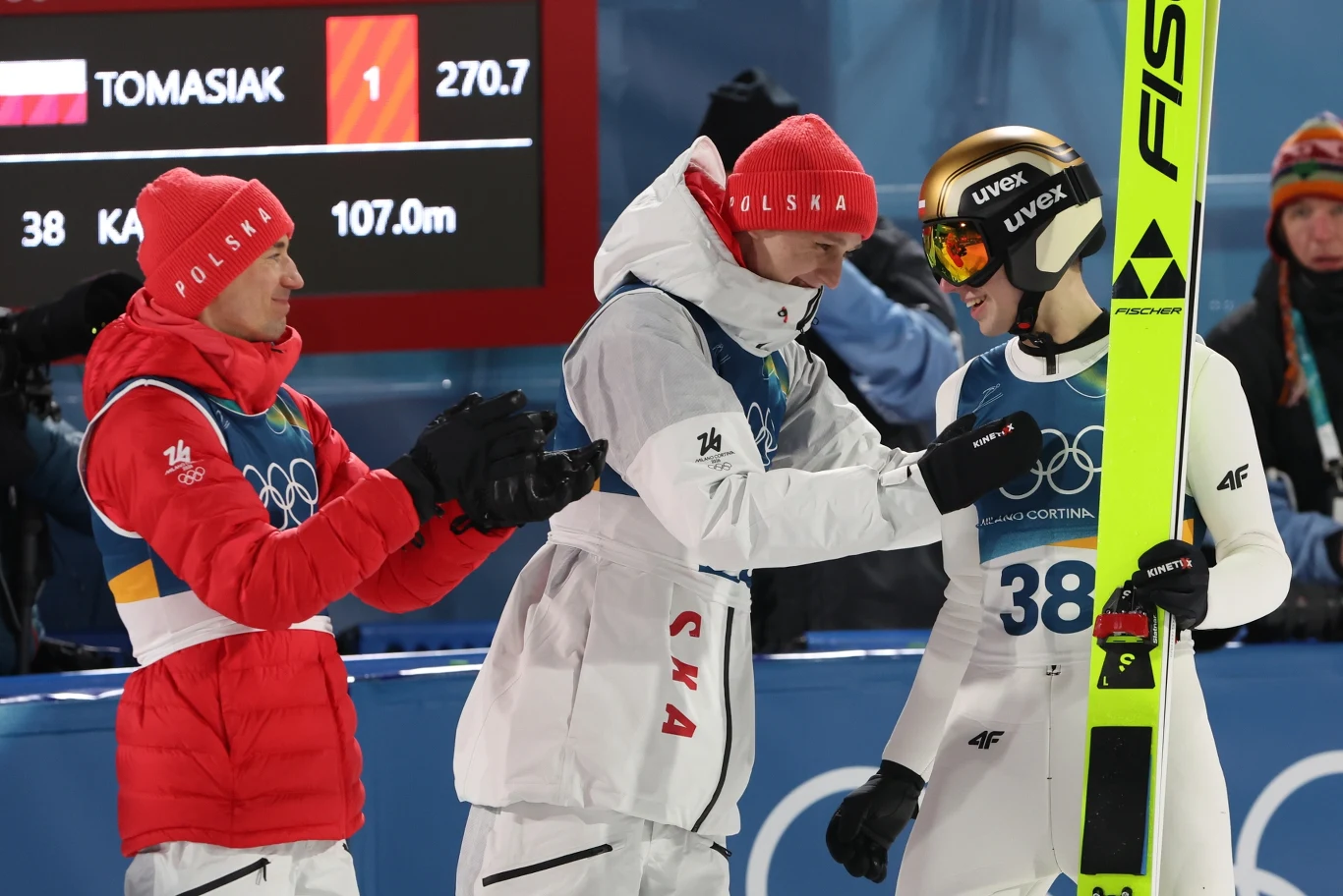 Od lewej: Kamil Stoch, Paweł Wąsek, Kacper Tomasiak Zawodnik w kombinezonie narciarskim oraz kasku z nartą w rękach stoi uśmiechnięty, otoczony przez dwóch członków drużyny w czerwonych i białych kurtkach z napisami Polska, trwa gratulacja po udanym skoku na tle tablicy z wynikami.