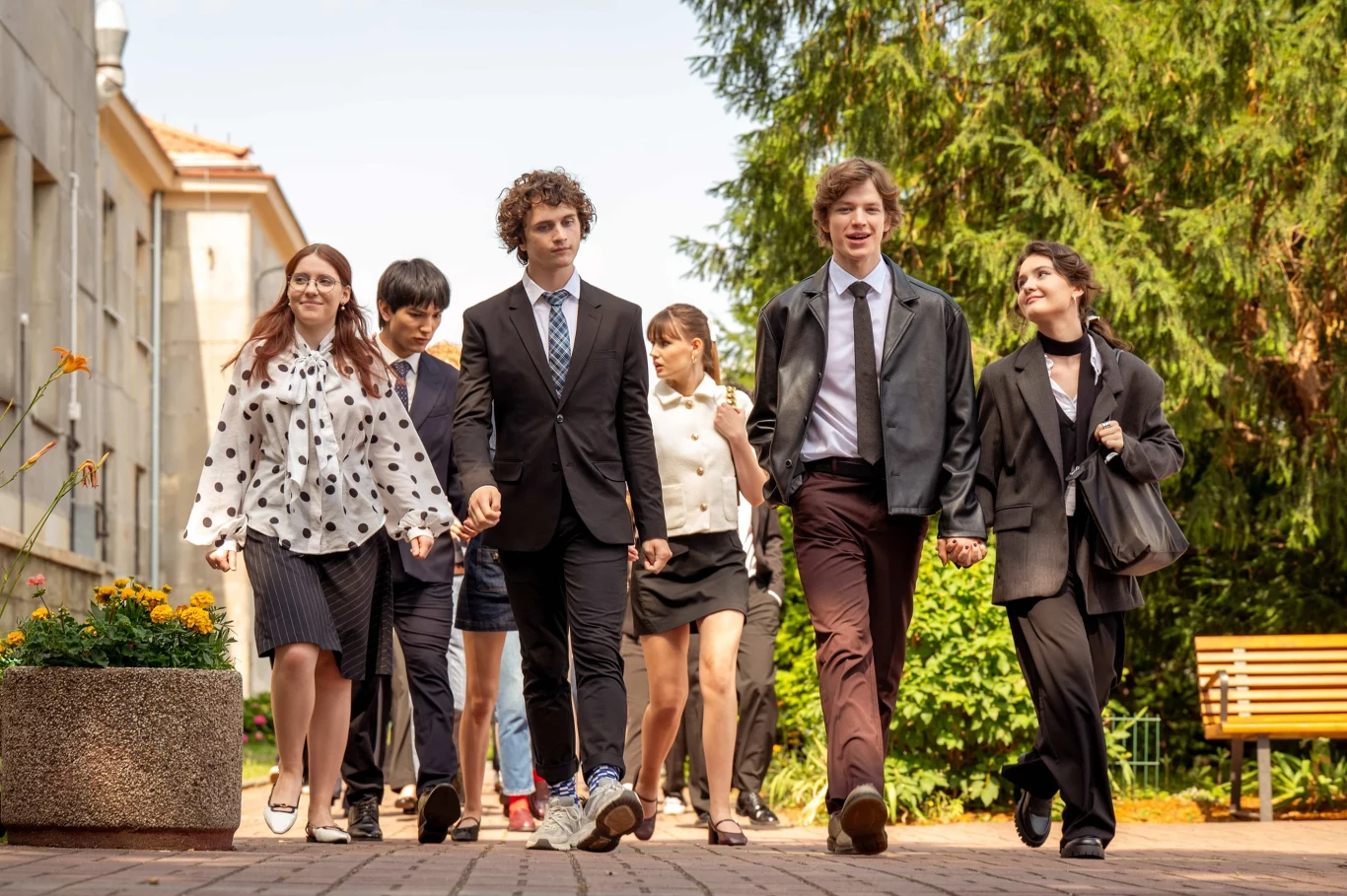 Grupa młodych osób ubranych elegancko idzie razem chodnikiem, otoczona zielenią i budynkiem szkolnym w tle, większość z nich uśmiechnięta, niektórzy trzymają się za ręce.