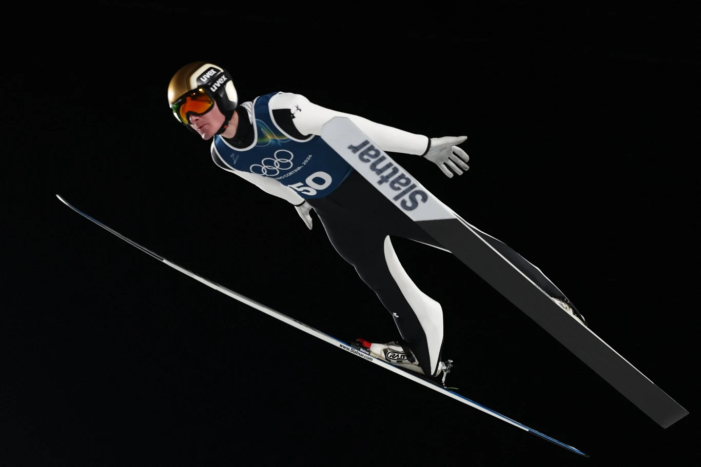 Domen Prevc Skoczek narciarski w locie podczas zawodów olimpijskich, ubrany w kombinezon startowy, kask i gogle, na tle ciemnego nieba.