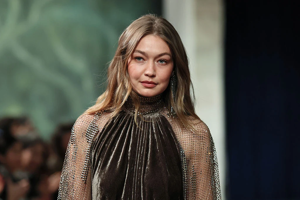 Gigi Hadid na wybiegu Ralph Lauren podczas New York Fashion Week Młoda kobieta o długich, jasnych włosach ubrana w elegancką, błyszczącą suknię z wysokim kołnierzem stoi na tle rozmytego tłumu oraz zielonej ściany.