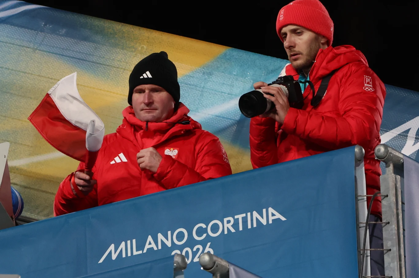 Dwaj mężczyźni ubrani w czerwone kurtki z logo reprezentacji Polski stoją na trybunie, jeden trzyma polską flagę, drugi aparat fotograficzny; w tle transparent z napisem Milano Cortina 2026.