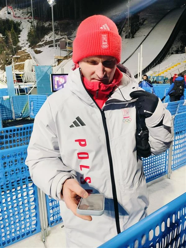 Maciej Maciusiak Mężczyzna ubrany w biały kombinezon z napisem 'Polska' oraz czerwonej czapce stoi na tle niebieskich barierek przy skoczni narciarskiej, trzyma w ręku telefon i radiostację.