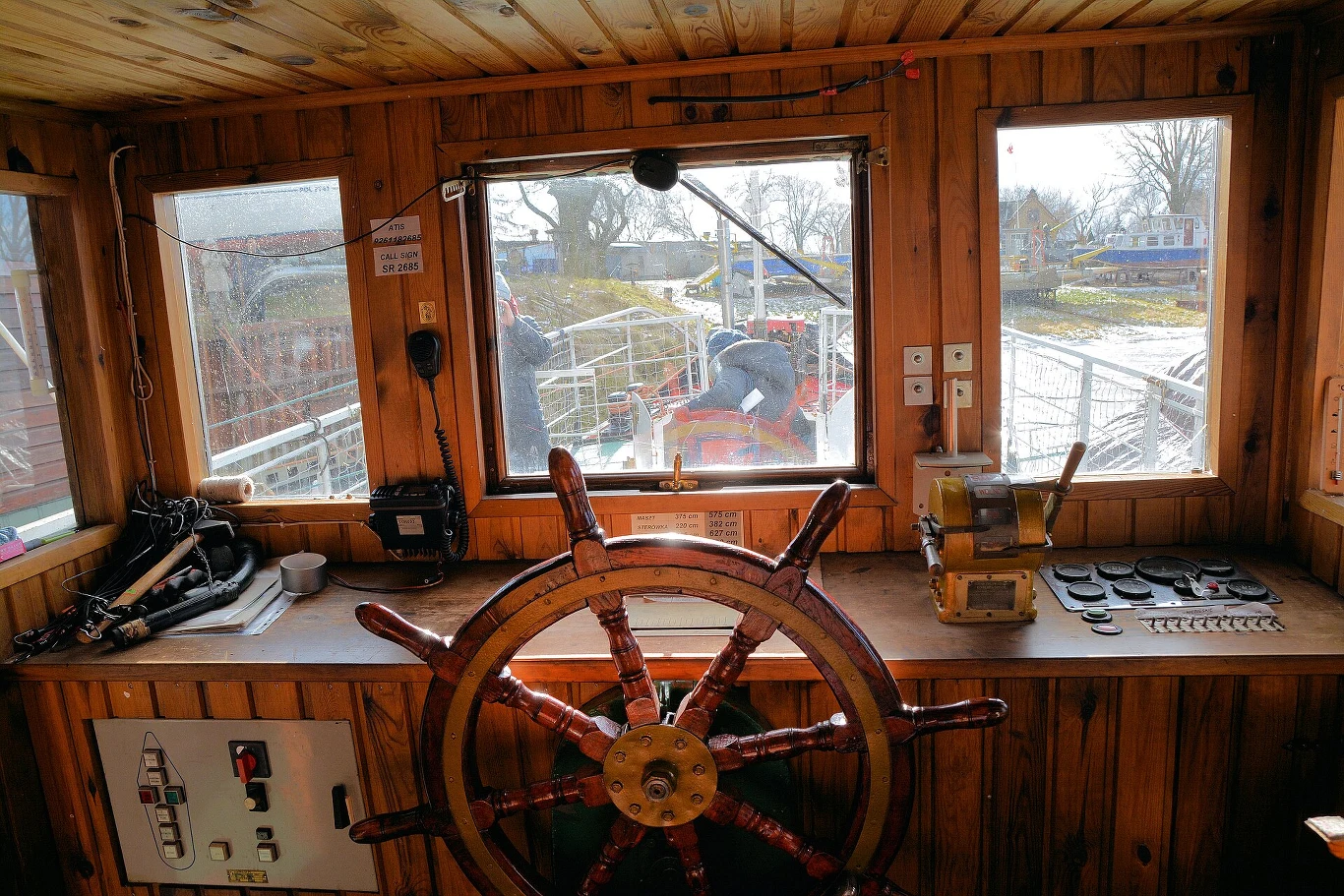 Drewniane wnętrze sterówki statku z dużym kołem sterowym na pierwszym planie, licznymi przyciskami i urządzeniami na blacie oraz widokiem na pokład i otoczenie przez okna.