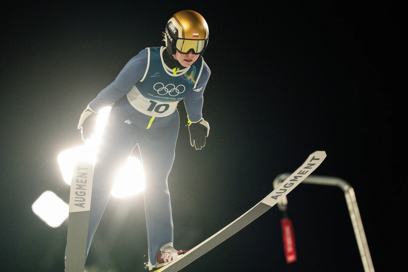 Skoki narciarskie. Pola Bełtowska w akcji Zawodnik w kombinezonie narciarskim i złotym kasku wykonuje skok na dużej skoczni narciarskiej podczas zawodów olimpijskich, w tle silne oświetlenie stadionowe oraz ciemne niebo.