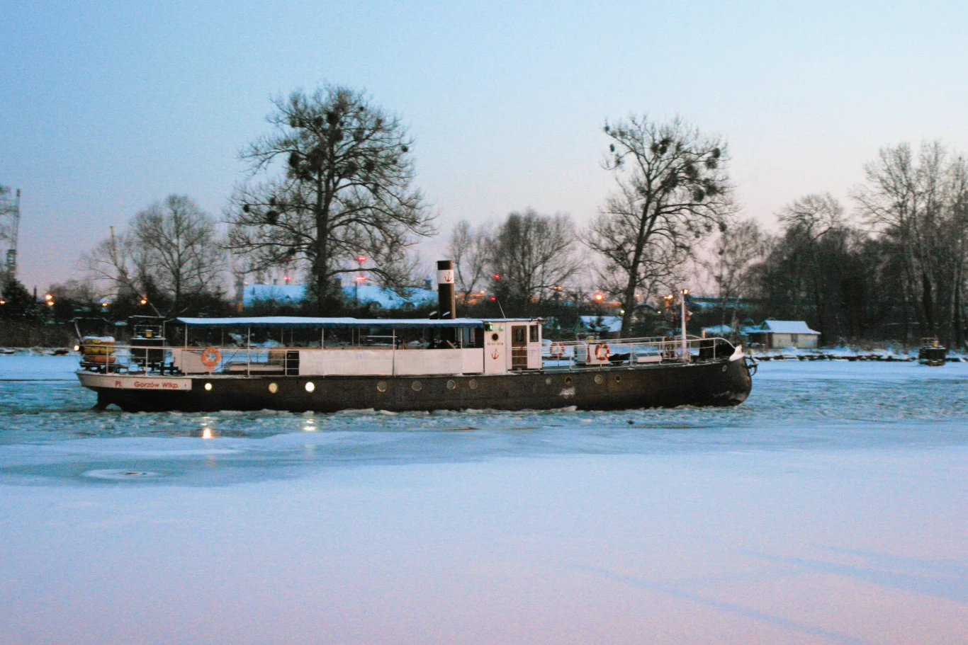 Statek poruszający się po zamarzniętej rzece, otoczony zimowym krajobrazem, w tle widoczne nagie drzewa i kilka budynków.