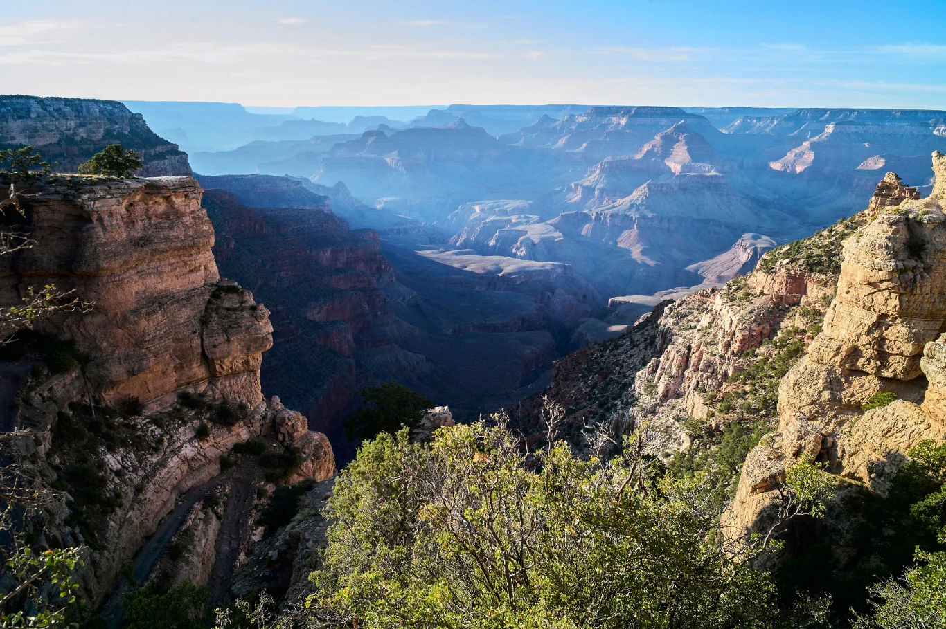 Grand Canyon, USA. Obcokrajowcy zapłacą więcej za wstęp do parków narodowych Rozległy widok skalistego kanionu z licznymi warstwami skał, roślinnością na pierwszym planie oraz rozproszonym światłem i mgłą unoszącą się nad głęboką doliną. Przestrzeń poprzecinana ostrymi krawędziami urwisk, w tle ukazują się kolejne linie wzniesi...