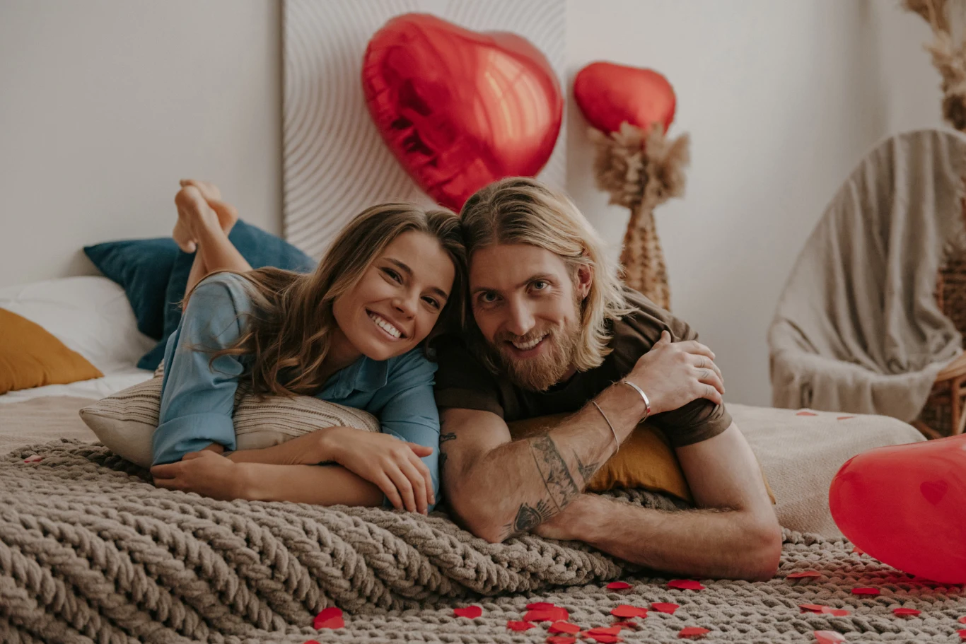 Uśmiechnięta para leży razem na łóżku pokrytym grubym, beżowym kocem, na tle dekoracji z czerwonymi balonami w kształcie serca i płatkami róż rozsypanymi na narzucie, tworząc romantyczną atmosferę.