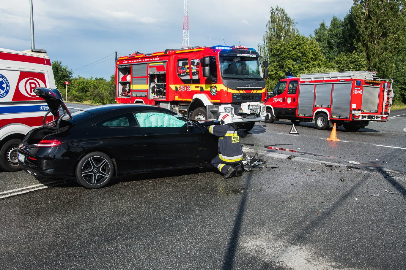 Wiele nowości w przepisach dotyczy straży pożarnej Czarny samochód osobowy po wypadku drogowym z widocznymi uszkodzeniami przodu, strażak klęczy obok pojazdu w trakcie prowadzenia akcji ratowniczej, w tle dwa wozy strażackie oraz ambulans, część drogi zabezpieczona pachołkami.