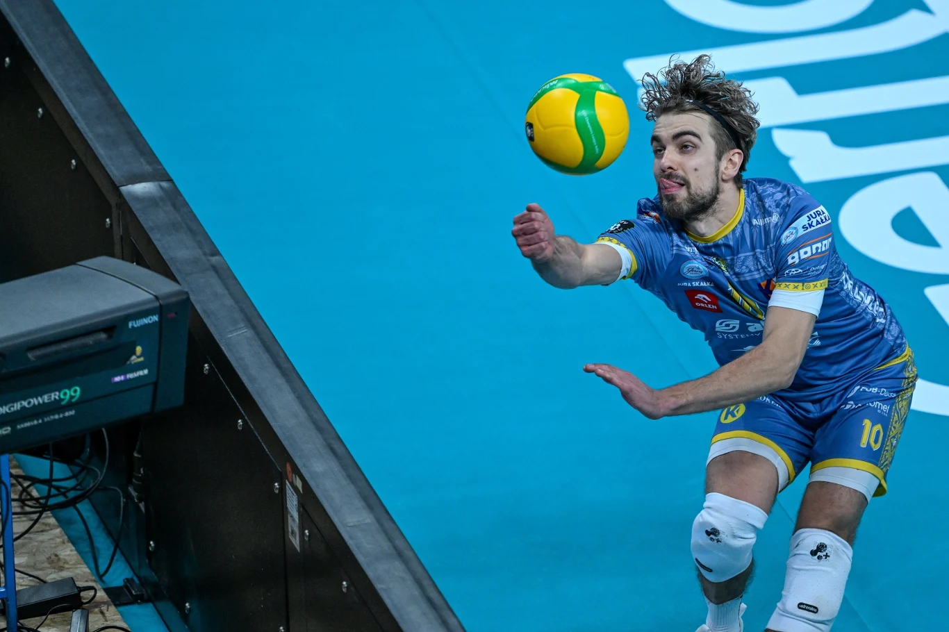 Siatkarz w niebieskim stroju drużynowym wykonuje dynamiczny ruch, próbując odbić piłkę podczas meczu na hali sportowej