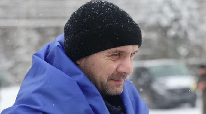 Mężczyzna w czapce i niebieskim okryciu stoi na śniegu, w tle rozmyta zimowa sceneria z niewyraźnymi elementami auta.