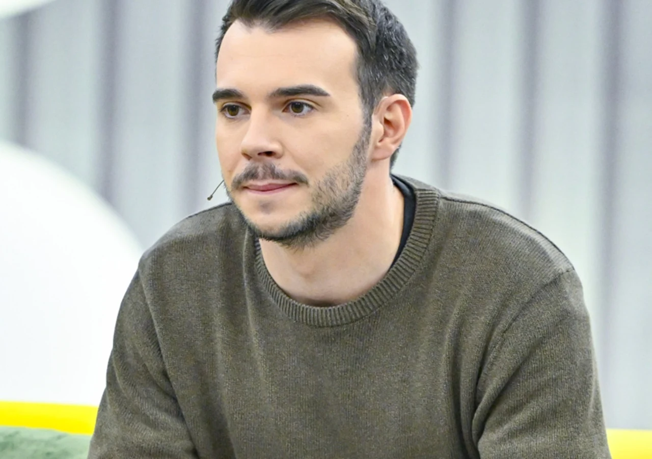 Albert Kosiński Młody mężczyzna z zarostem w brązowym swetrze siedzi przed jasnoszarym, rozmytym tłem, na twarzy delikatny uśmiech, na uchu mikrofon słuchawkowy.