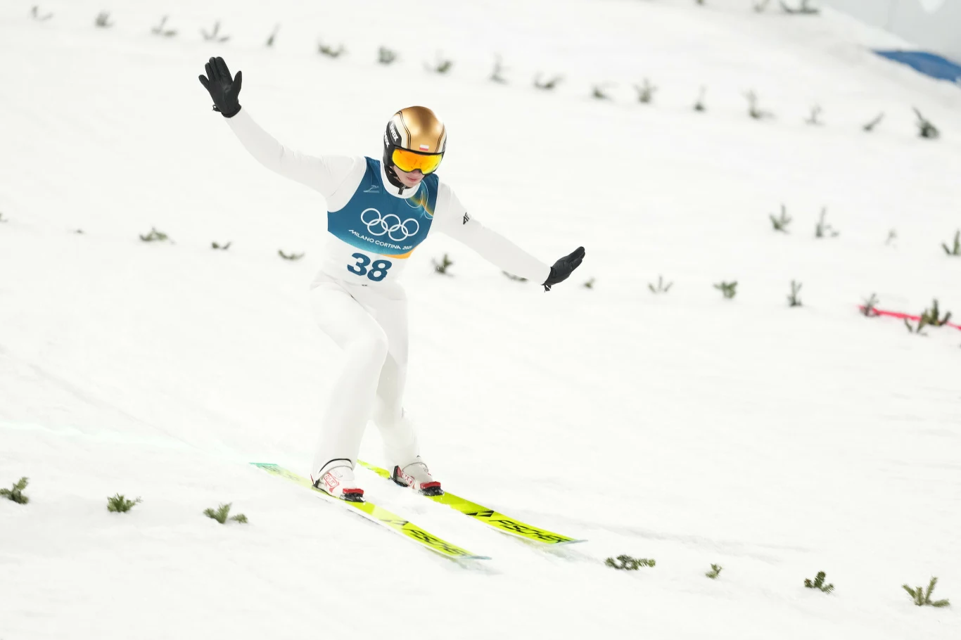 Kacper Tomasiak Sportowiec w kombinezonie narciarskim, z kaskiem i okularami, ląduje na nartach podczas konkursu skoków narciarskich na zimowych igrzyskach olimpijskich, unosi ręce dla zachowania równowagi na śnieżnym stoku.