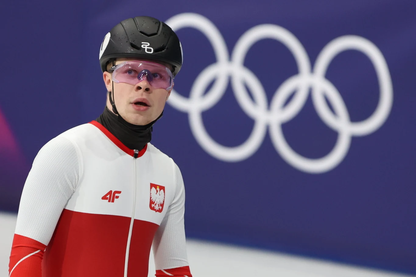 Polski panczenista w sportowym stroju, kasku i okularach ochronnych na tle symbolu igrzysk olimpijskich.