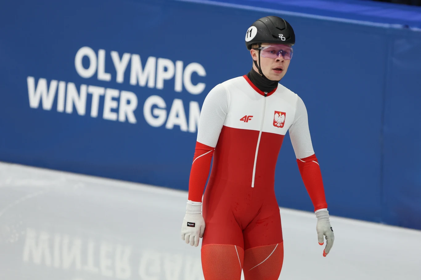 Zawodnik w czerwono-białym stroju z herbem Polski na piersi stoi na torze lodowym podczas Zimowych Igrzysk Olimpijskich, na głowie kask z numerem, w tle niebieska plansza z napisem Olympic Winter Games.