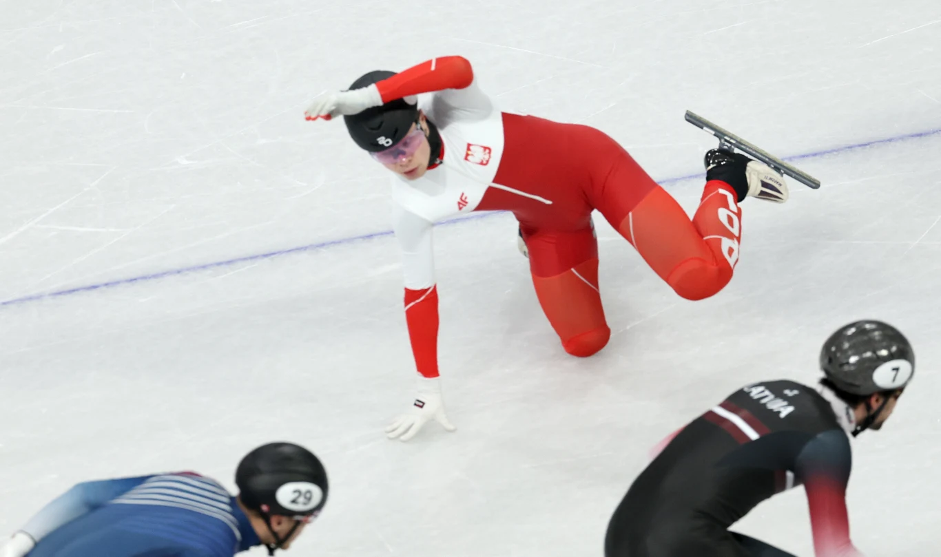 Łyżwiarz w biało-czerwonym stroju upada na lodowisku podczas wyścigu, w tle dwóch innych zawodników w trakcie rywalizacji.