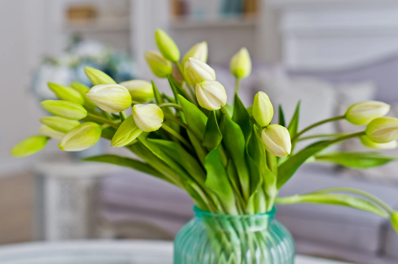 Bukiet białych tulipanów w zielonym wazonie ustawiony na stole w jasnym, eleganckim wnętrzu.