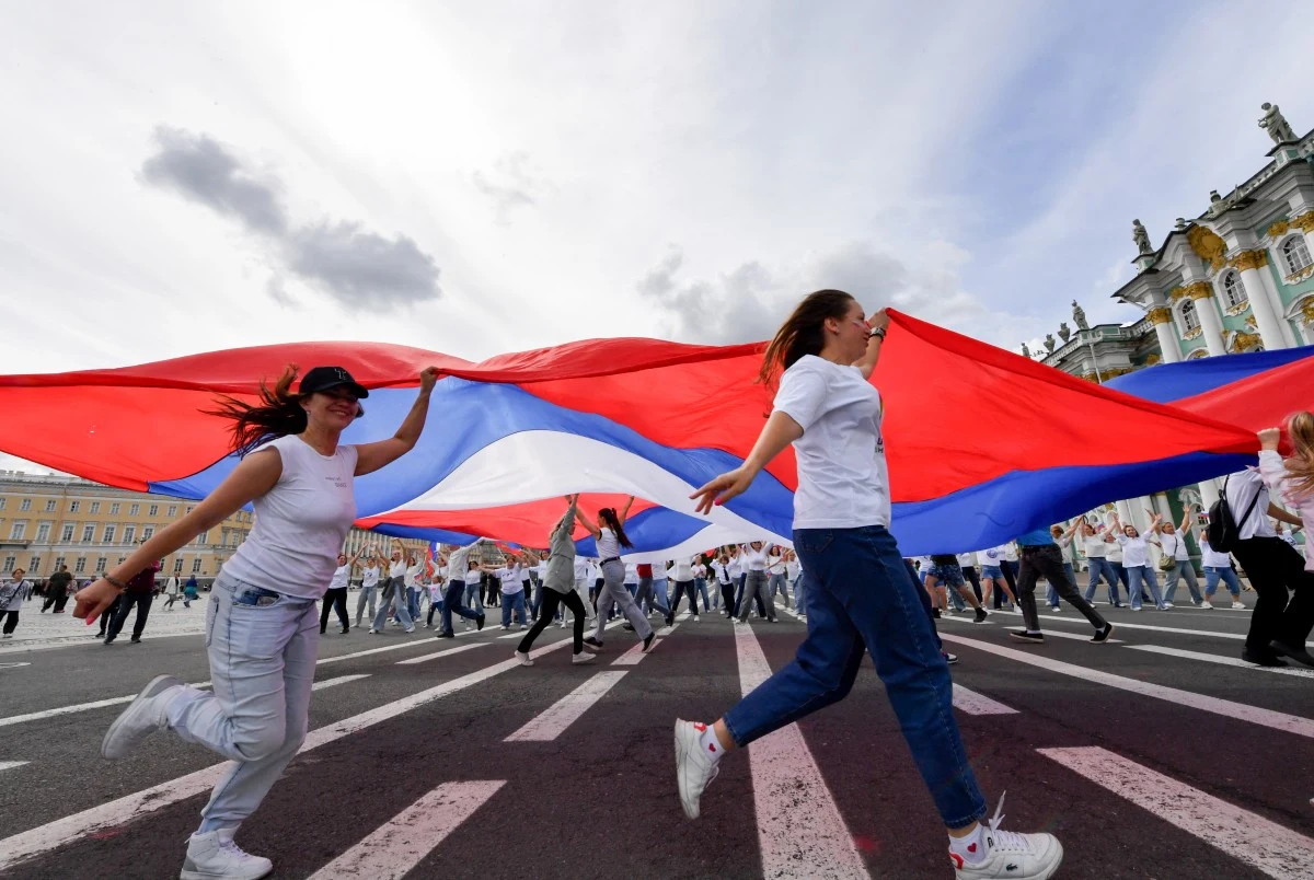 Uczestnicy maszerują pod rozpiętą ogromną flagą w barwach biało-niebiesko-czerwonych, ubrani głównie na biało, tworząc efektowną kompozycję na tle szerokiej ulicy i reprezentacyjnych budynków.