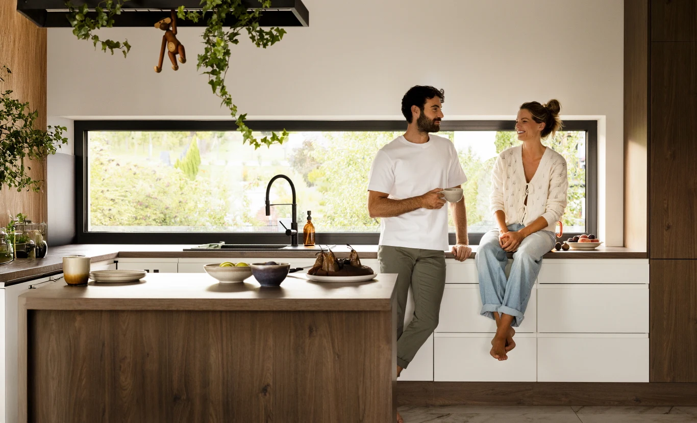 Bilans energetyczny okien nie dotyczy wyłącznie sezonu grzewczego. Latem problem odwraca się — zamiast ucieczki ciepła mamy nadmierne zyski energii słonecznej przez przeszklenia Para rozmawiająca w nowoczesnej, jasnej kuchni z dużym oknem, otoczona roślinami i eleganckimi akcesoriami kuchennymi, kobieta siedzi na blacie, mężczyzna stoi obok niej trzymając kubek.