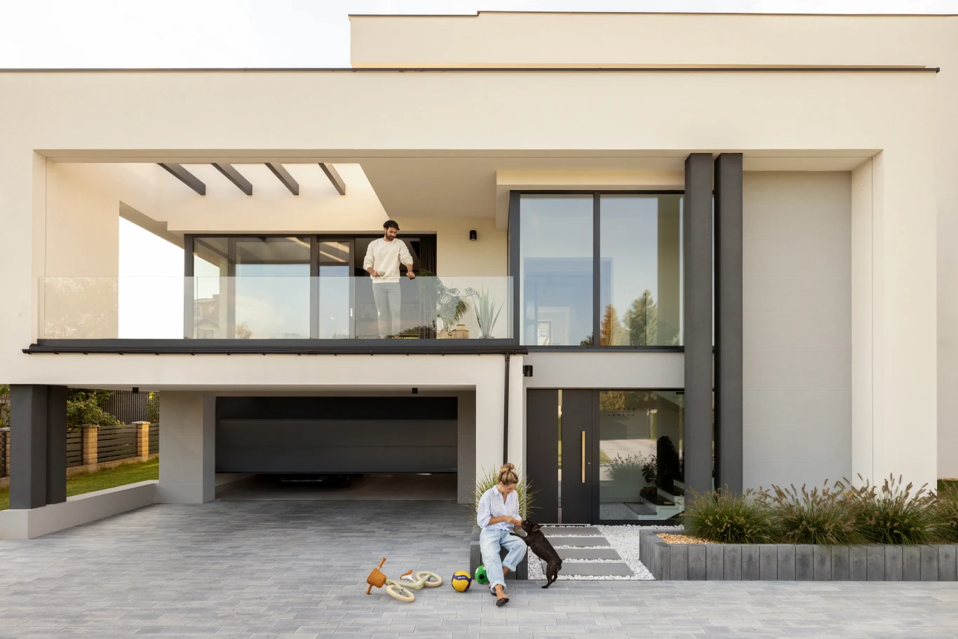 Świadomy wybór wymaga spojrzenia systemowego - nie na jedną liczbę z reklamy, lecz na komplet parametrów wpływających na komfort i koszty przez cały rok Nowoczesny dwupiętrowy dom z dużymi przeszkleniami, na tarasie stoi mężczyzna, przed wejściem kobieta siedzi na ziemi obok psa i zabawek, przestronny podjazd oraz zadbana roślinność wokół budynku.