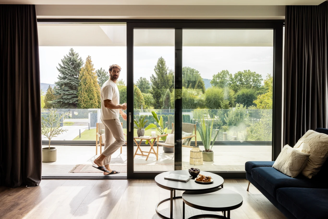 Kluczowe jest sprawdzenie, czy deklarowany parametr dotyczy standardowego, realnie zamawianego wymiaru okna, czy wyłącznie najmniejszej konfiguracji referencyjnej Mężczyzna w jasnych ubraniach stoi w drzwiach tarasowych nowoczesnego salonu, wychodząc na balkon z widokiem na zielony ogród i drzewa. Na balkonie ustawione są rośliny doniczkowe oraz stolik z krzesłami, wnętrze salonu cechuje się jasnością i minimali...