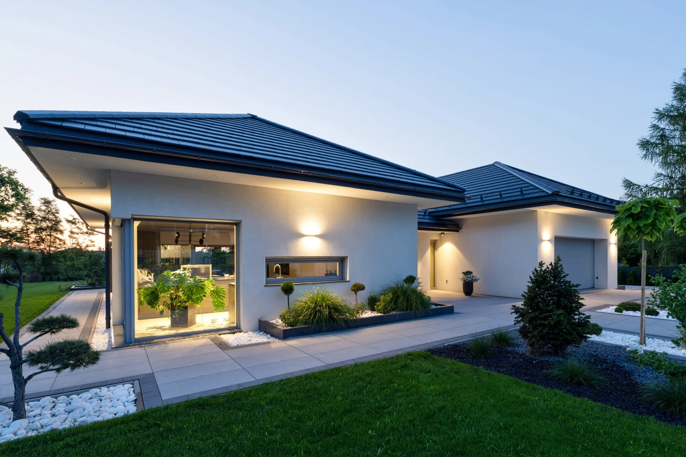 Coraz więcej producentów projektuje stolarkę jako element większego systemu domu. W ofercie WIŚNIOWSKI obejmuje to zarówno okna PVC (PRIMO, DEVELO), jak i rozwiązania aluminiowe Nowoczesny dom jednorodzinny z jasną elewacją, dużymi oknami i starannie utrzymanym ogrodem. Oświetlenie zewnętrzne subtelnie podkreśla linię budynku, a na tarasie widoczne są ozdobne rośliny w donicach i przycinane krzewy. Otoczenie domu porządkuje tr...