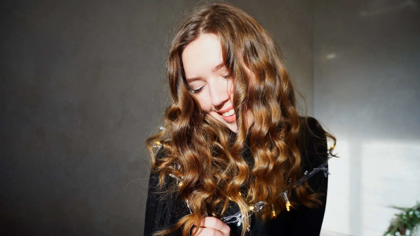 Młoda kobieta z długimi, falowanymi włosami i uśmiechem na twarzy, ubrana w ciemny strój, lekko pochylona głową, w tle neutralna, jasna ściana.