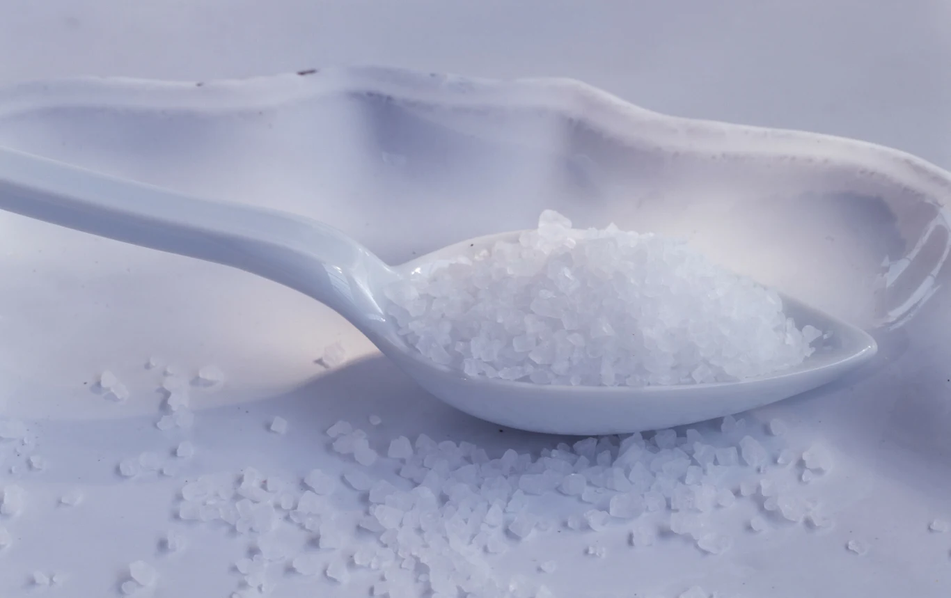 Biała sól morska rozsypana na płaskiej powierzchni oraz umieszczona na białej porcelanowej łyżeczce.