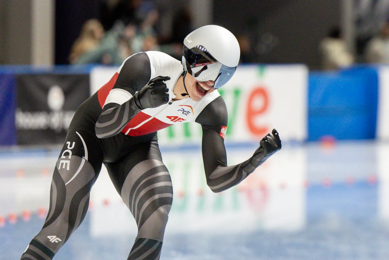 Zawodnik w profesjonalnym stroju łyżwiarskim świętuje sukces na lodowisku, zaciśnięte pięści wyrażają emocje po osiągnięciu celu. Tło rozmyte, skupienie na sportowcu.