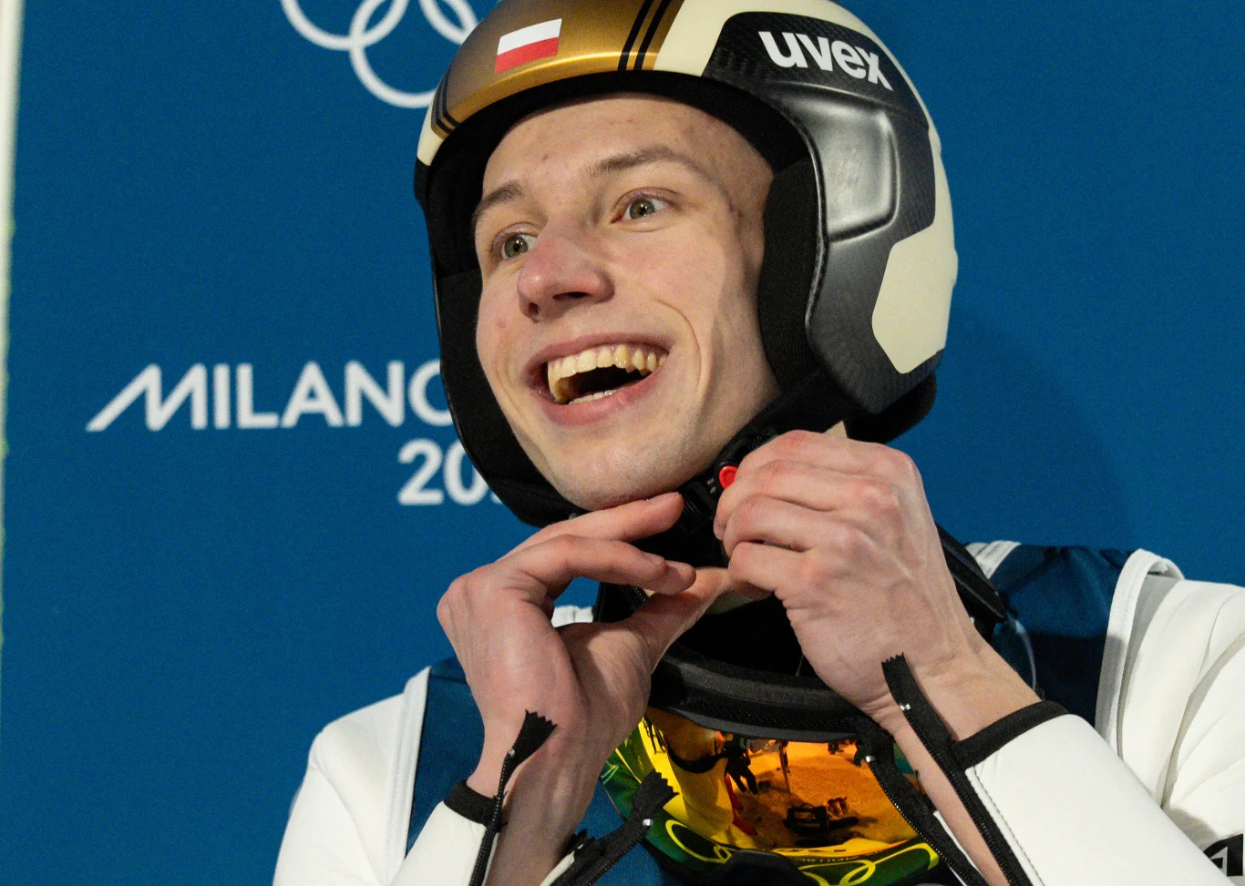 Skoczek narciarski w kasku i kombinezonie sportowym uśmiecha się szeroko, przygotowując się do odpięcia paska przy kasku, na tle napisu Milan oraz symbolu olimpijskiego.