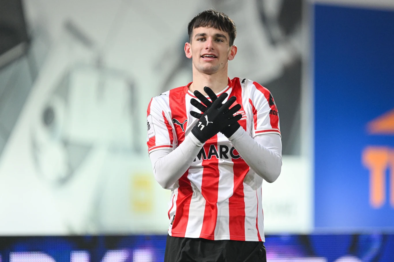 Młody piłkarz ubrany w biało-czerwony pasiasty strój sportowy stoi na boisku i wykonuje gest rękami ułożonymi na klatce piersiowej, wokół niego widoczna rozmyta arena sportowa.