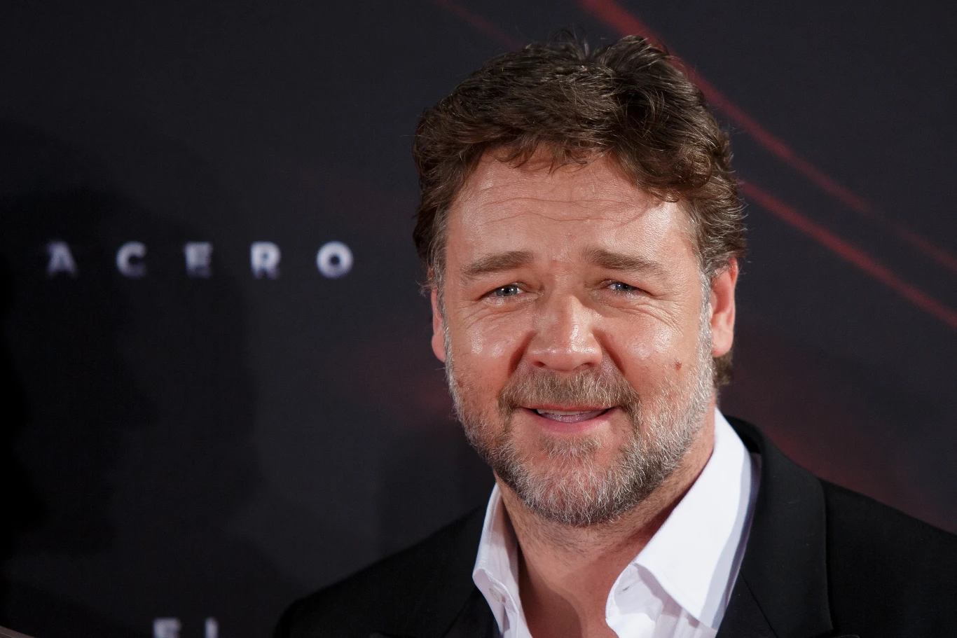 Russell Crowe Mężczyzna w średnim wieku w eleganckim stroju, z krótką brodą i uśmiechem, stoi na tle ciemnej ścianki z niewyraźnymi napisami.