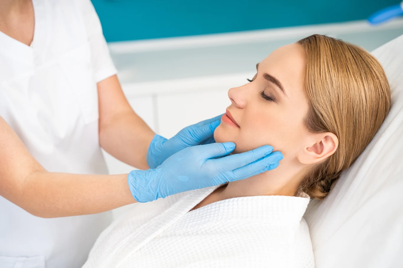 Specjalistka w białym uniformie i niebieskich rękawiczkach sprawdza stan skóry twarzy młodej kobiety siedzącej na fotelu zabiegowym, kobieta jest zrelaksowana i ma zamknięte oczy.
