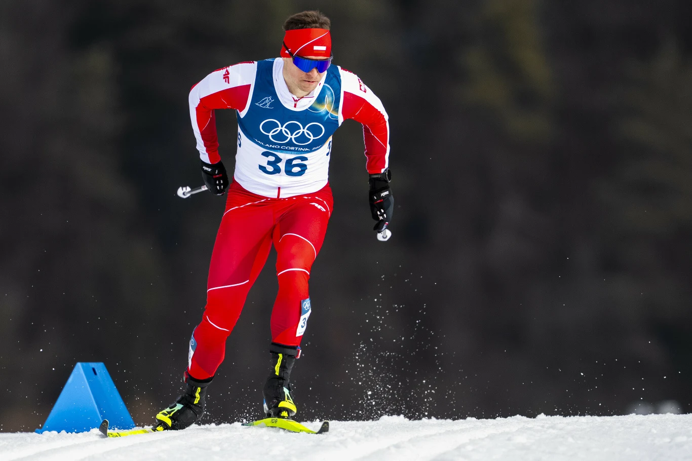 Biegacz narciarski w czerwonym stroju olimpijskim z numerem 36, energicznie pokonujący trasę na tle śnieżnej scenerii, w dłoniach trzyma kijki, za nim widoczne ciemne drzewa.