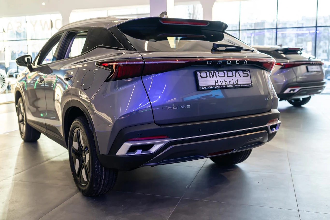 Nowoczesny samochód SUV marki OMODA S Hybrid w kolorze szarym, prezentowany w salonie samochodowym pod oświetleniem, widok od tyłu, dobrze widoczne detale lamp i tabliczka rejestracyjna.
