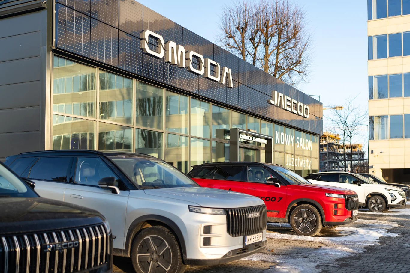 Kilka nowoczesnych samochodów osobowych ustawionych przed przeszklonym budynkiem salonu samochodowego z widocznymi szyldami marek Omoda oraz Jaecoo; zimowa aura i odbicia budynków w szybach.