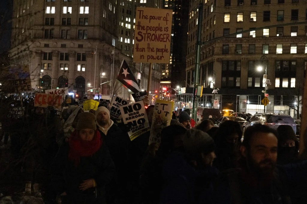 Protest przeciwko działalności ICE Tłum ludzi podczas nocnej demonstracji w miejskiej scenerii, wśród nich widoczne są transparenty z hasłami politycznymi oraz flagi, otaczające oświetlone budynki w tle.